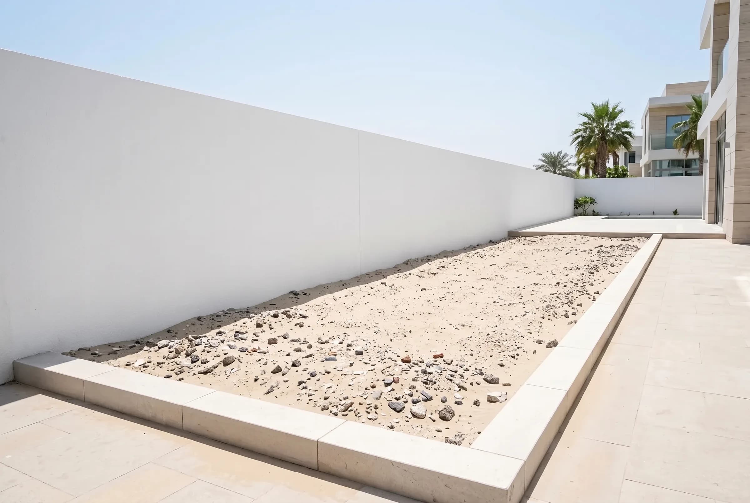 Before: a bare new villa garden bed of pale compacted sand and builders' rubble along a white wall in Dubai
