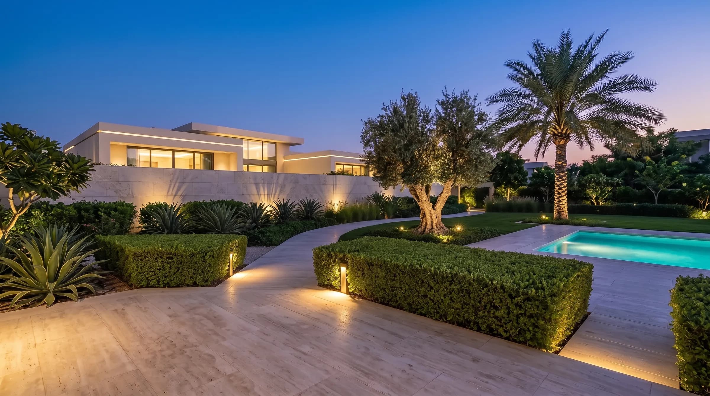 Luxury Dubai villa garden hero at blue hour: layered warm lighting on a sculptural olive tree, clipped hedges and travertine stone by a lit pool