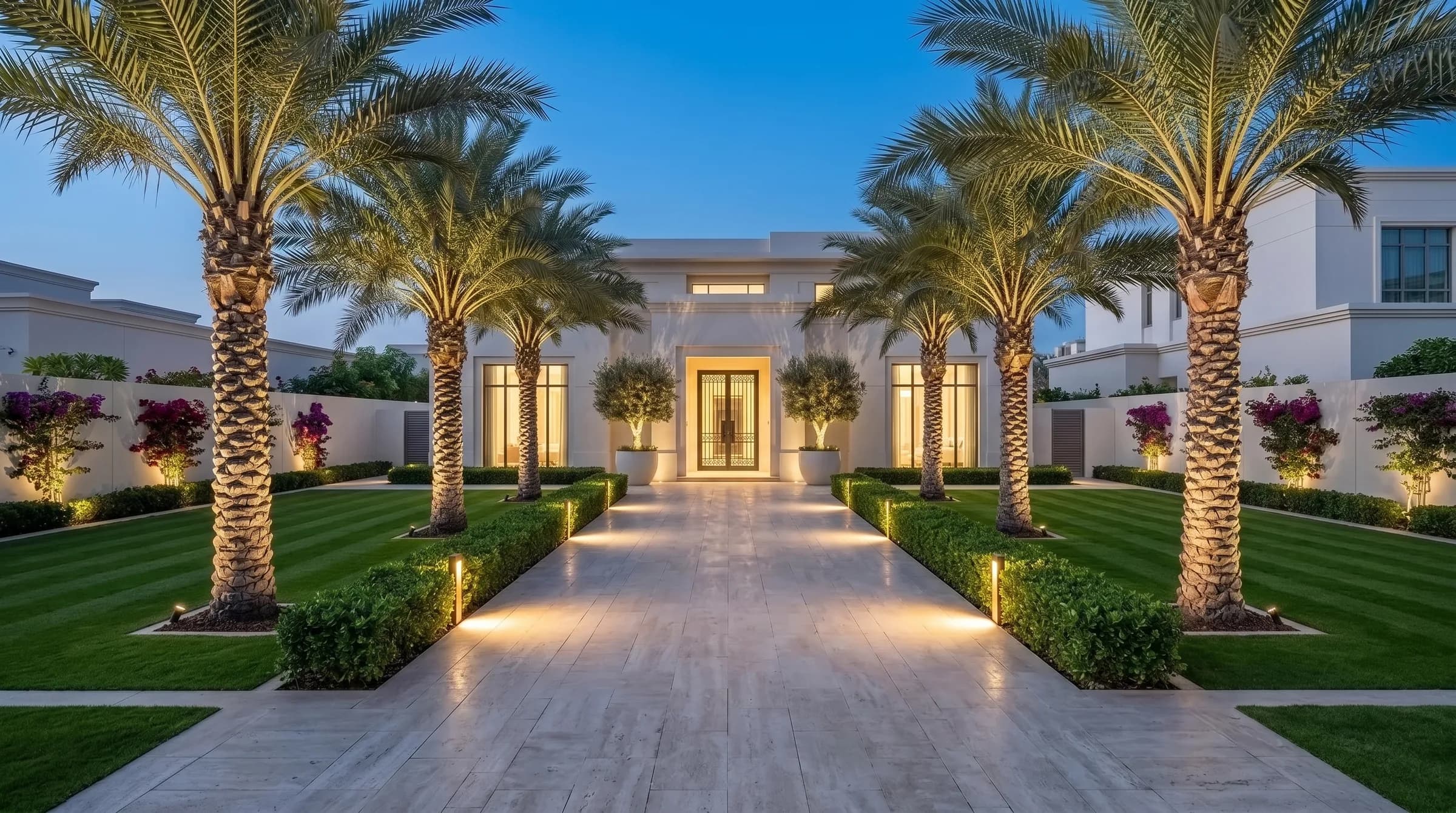 Manicured Dubai villa garden at blue hour: uplit date palm allee, softly lit hedges and a glowing entrance at the end of the travertine path