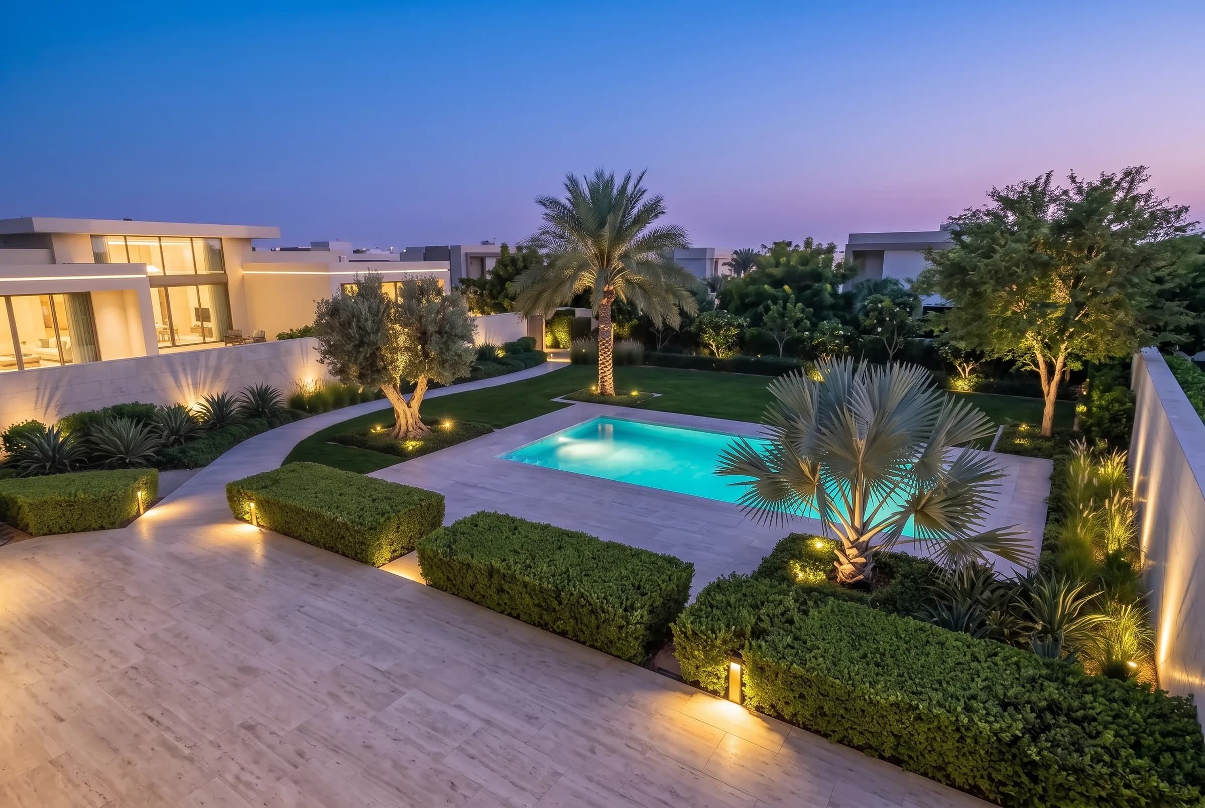 Elevated dusk overview of a full luxury Dubai villa garden: lit stone terrace, pool and clipped hedges around uplit palms and an olive specimen
